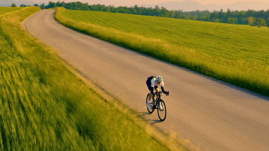A cyclist rides a road bike along a winding country road bordered by bright green fields under a warm golden sky.